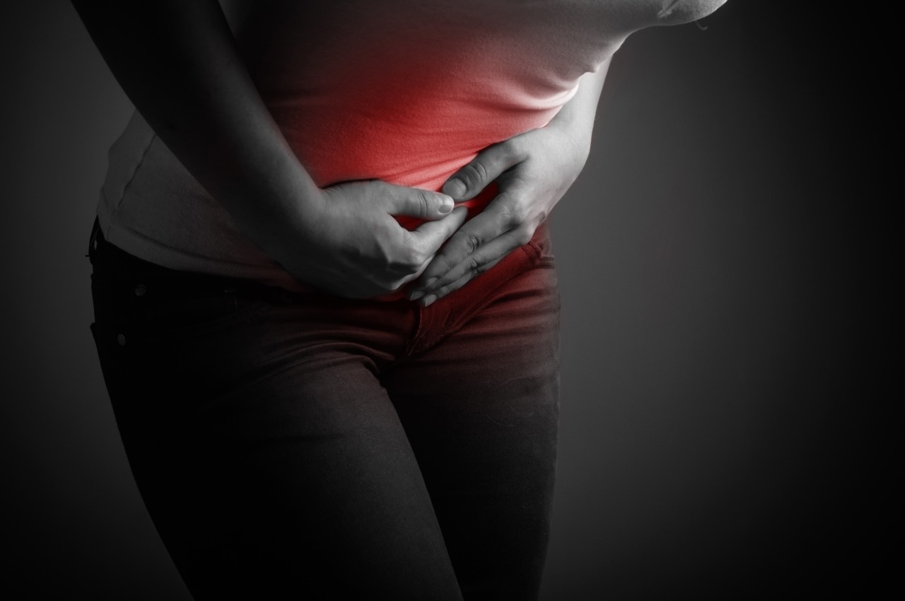 black and white close up of a woman's abdomen and thighs. She is wearing a white t-shirt and dark jeans, with her hands clutching her lower abdomen in pain. There is a red glow over the affected area.