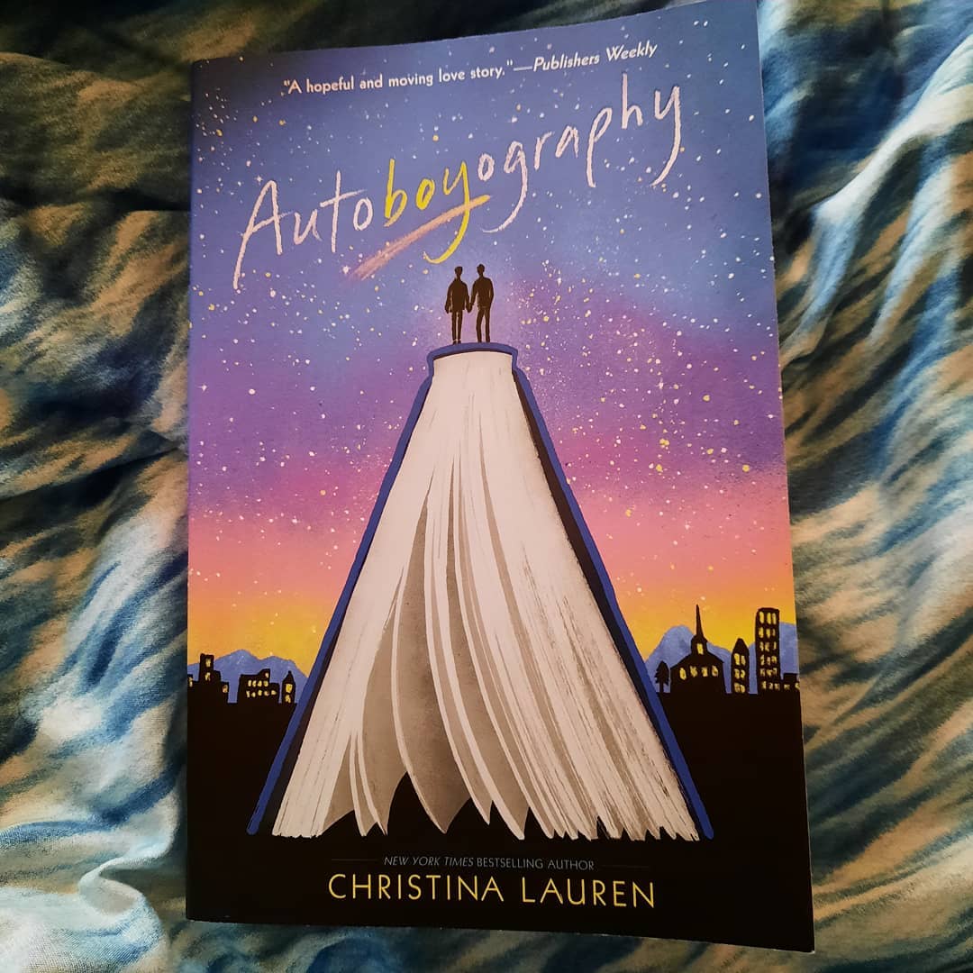 The book Autoboyography by Christina Lauren. The cover shows two silhouettes holding hands and standing on a giant book overlooking a city skyline at night.