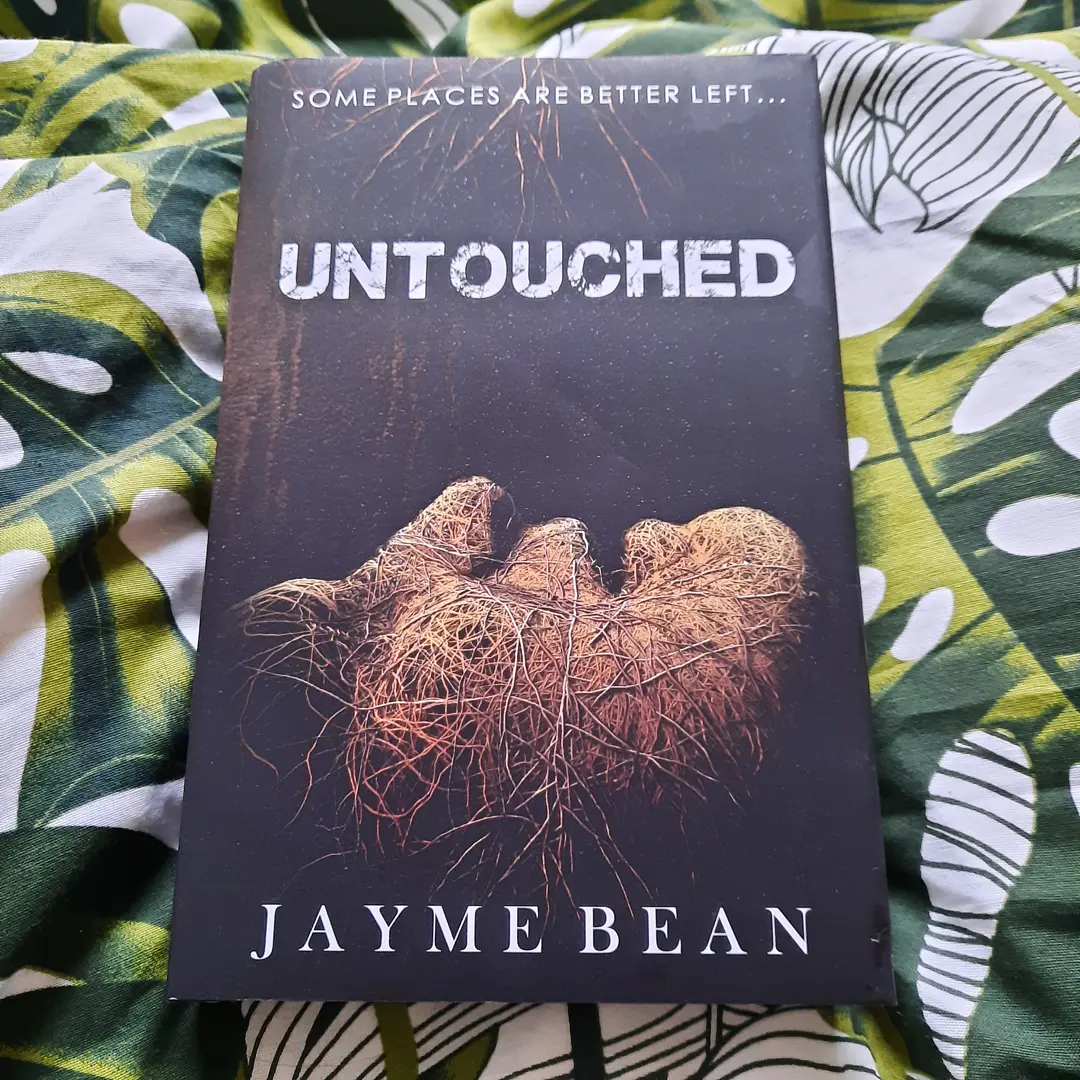 A hardback copy of the book Untouched by Jayme Bean, which is a dark brown cover with a face on it covered in vines and the words "some places are better left untouched" in white font