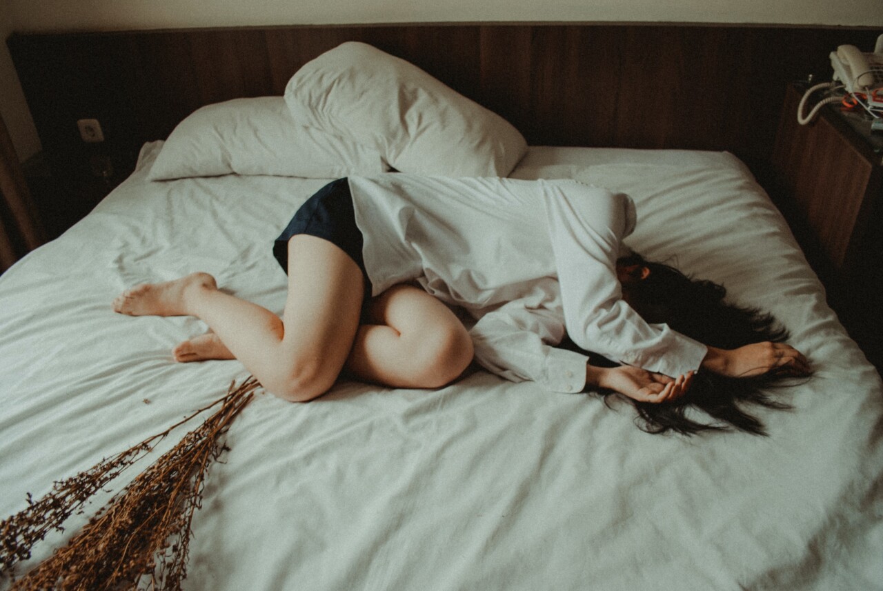 A white woman with long dark hair, is sprawled across an unmade bed. She is wearing a white shirt and black shorts and covering her face with her arms. It is a vision of despair, the best I could find to express the trauma of PIP